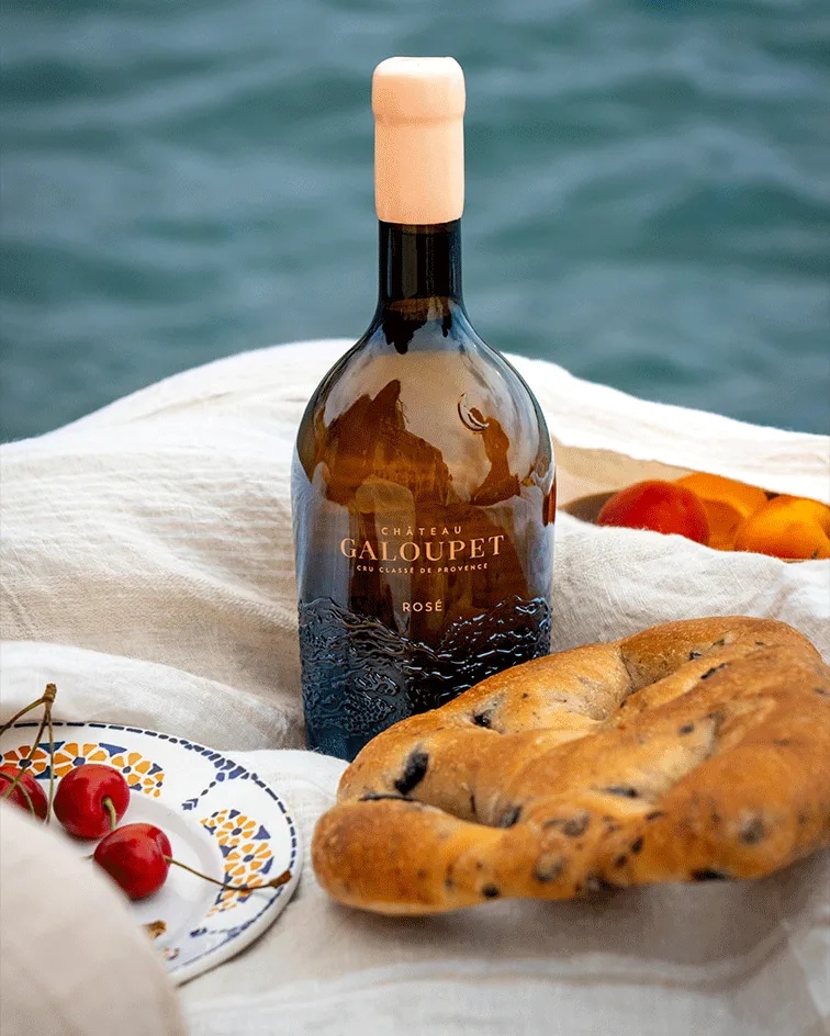 Bottle of Château Galoupet Cru Classé Côtes de Provence Rosé on a rock over the Mediterranean sea, surronded by fruits and bread.